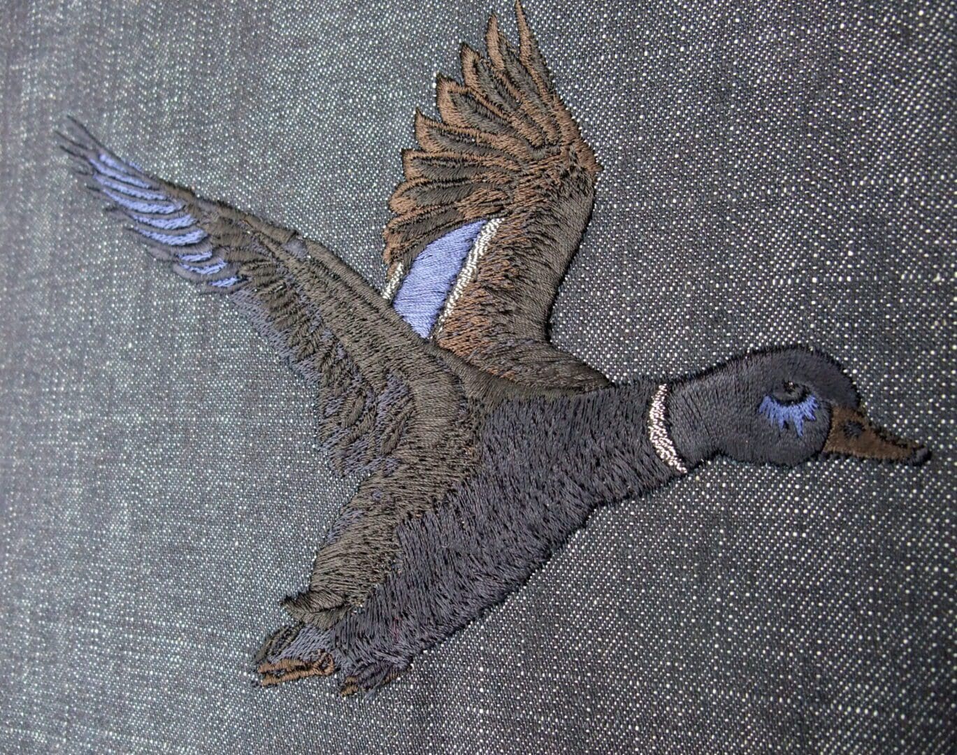 Nearly all black shade of embroidery for a flying Mallard duck design in stitch on black denim fabric