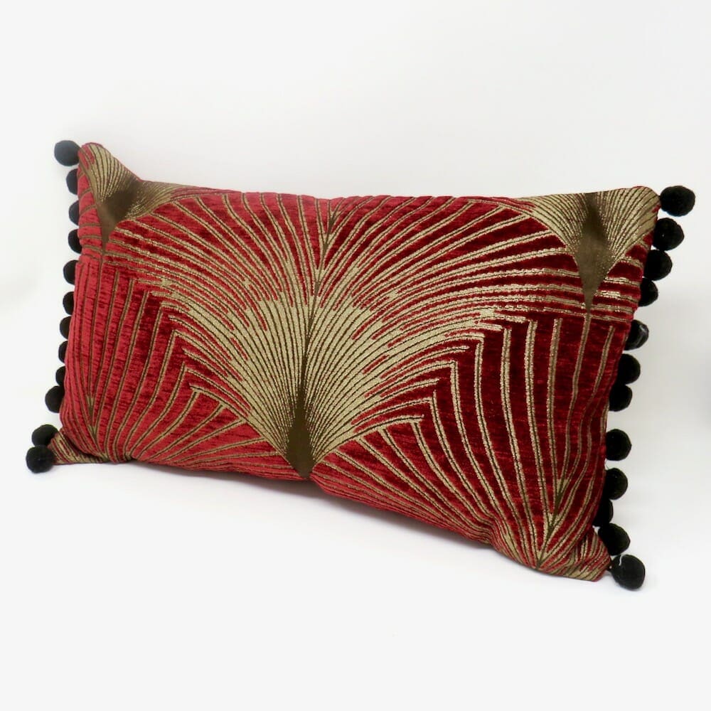 A burgundy and gold art deco style velvet cushion with a black pom pom trim.