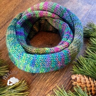 green and pink hand knitted double loop infinity scarf in vegan yarn, laying on wooden surface surrounded by pine fir boughs