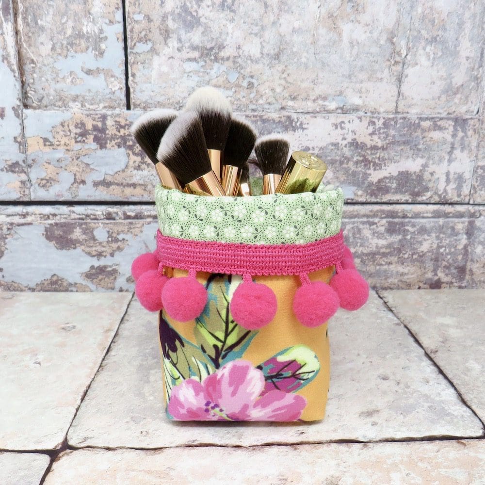 A mini velvet storage pot in a yellow pineapple print and with a cerise pom pom trim.