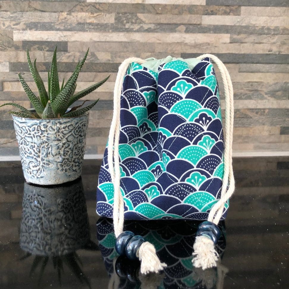 Mini drawstring bag in stylistic Japanese wave design in blue and teal with pale green lining, closing with two chunky shiny blue ceramic beads, standing on a black surface with small pot plant to the left