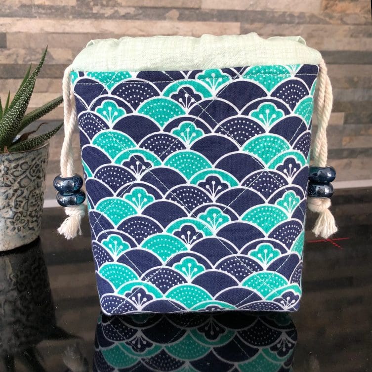 Mini drawstring bag in stylistic Japanese wave design in blue and teal with pale green lining, closing with two chunky shiny blue ceramic beads, standing on a black surface with small pot plant to the left