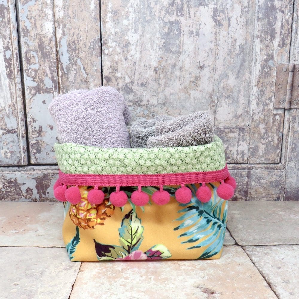 Velvet storage tub in a yellow pineapple print and with a cerise pom pom trim.