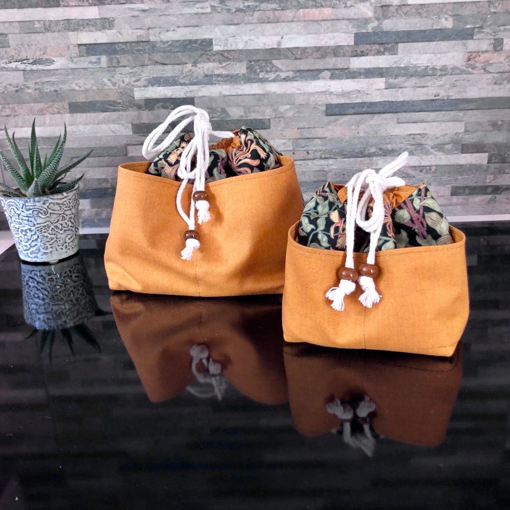 Set of large and small WeeBraw, drawstring bags in William Morris' Honeysuckle design on a black background, full width external ochre yellow pockets. Closing with natural unbleached cotton secured with chunky ceramic ochre beads on a glossy black surface, the bags are pulled closed and tied and sit on a shiny black surface.