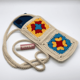 a bag made from 4 granny squares joined together to make a bag. sticking out of the top is a mobile phone and by the side is a tube of lip salve, the long thin handle is curled round the edge of the bag