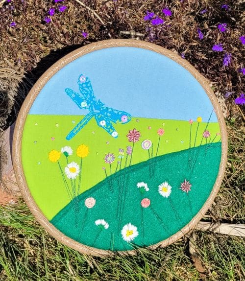 Hand embroidered 8 inch hoop of a dragonfly flying amongst flowers.