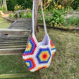 a crochet bag in the shape of a hexagon is hanging on a wooden bench. The bag is made up of hexagons in rainbow colours and joined with cream. The handle is cream and there is wooden hexagonal button at eavh end where it joins the bag