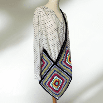 it's a crochet bag made of 4 squares to make a diamond shape. The squares are multicoloured rounds and are edged in black. The handle is black too. It is displayed on a mannequin. The background is white