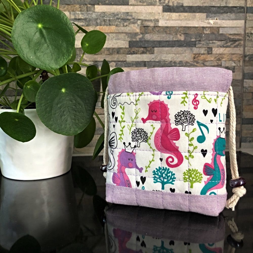 Mini drawstring bag in cute underwater design including pink and lilac seahorses with pale lilac base and lining, closing with two chunky shiny purple ceramic beads, standing on a black surface with small pot plant to the left