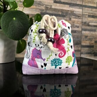 Closed and tied view of mini drawstring bag in cute underwater design including pink and lilac seahorses with pale lilac base and lining, closing with two chunky shiny purple ceramic beads, standing on a black surface with small pot plant to the left