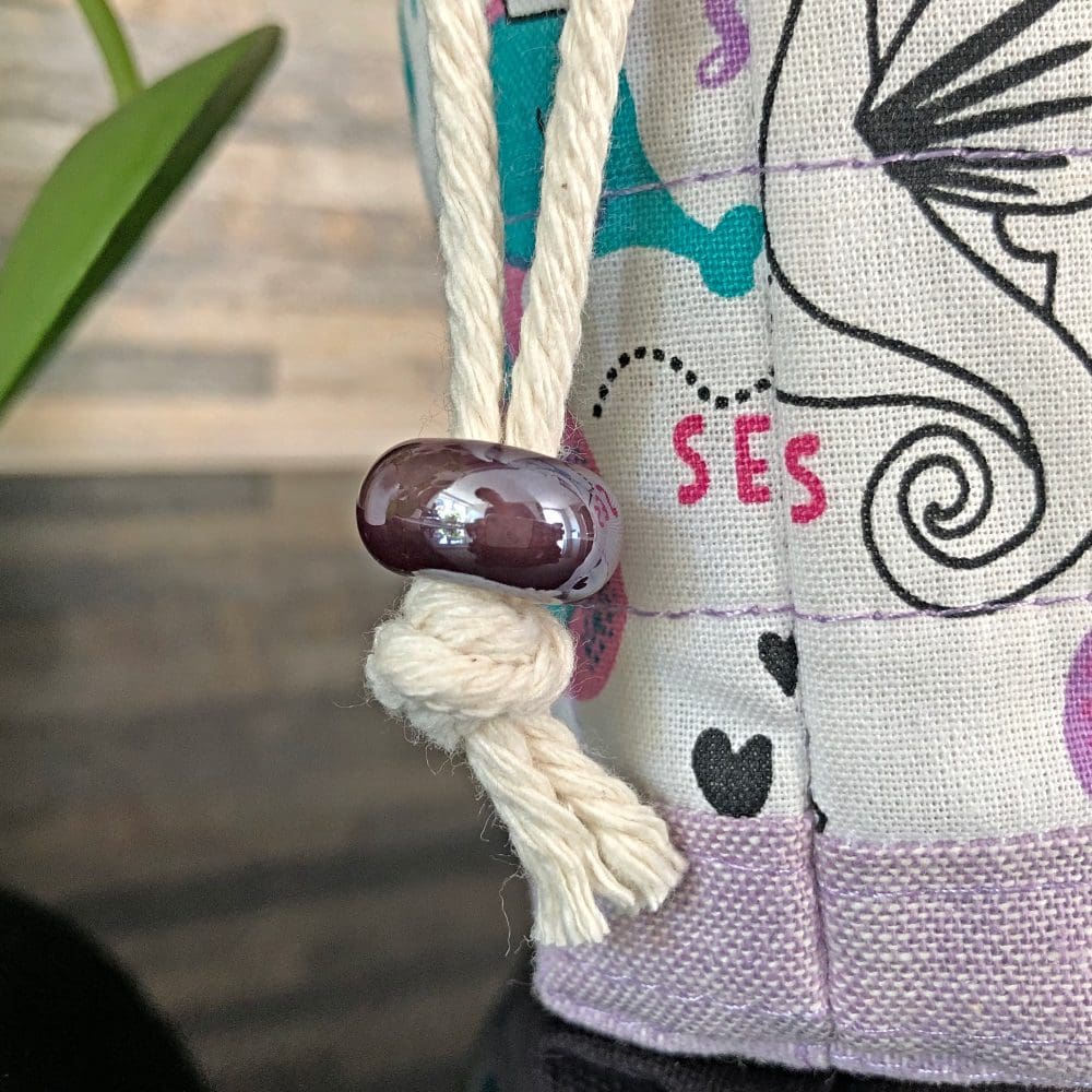 Close up view of the ceramic bead on the mini drawstring bag in cute underwater design including pink and lilac seahorses with pale lilac base and lining, closing with two chunky shiny purple ceramic beads, standing on a black surface with small pot plant to the left
