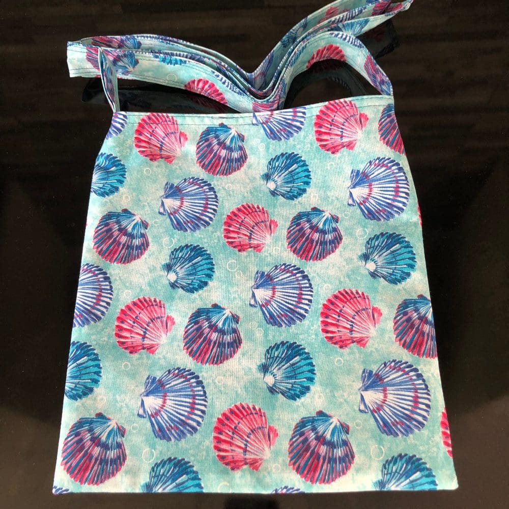 Adjustable cross body style mastectomy drain bag in pale blue with a pink and blue shell design and blue lining, on a shiny black surface.