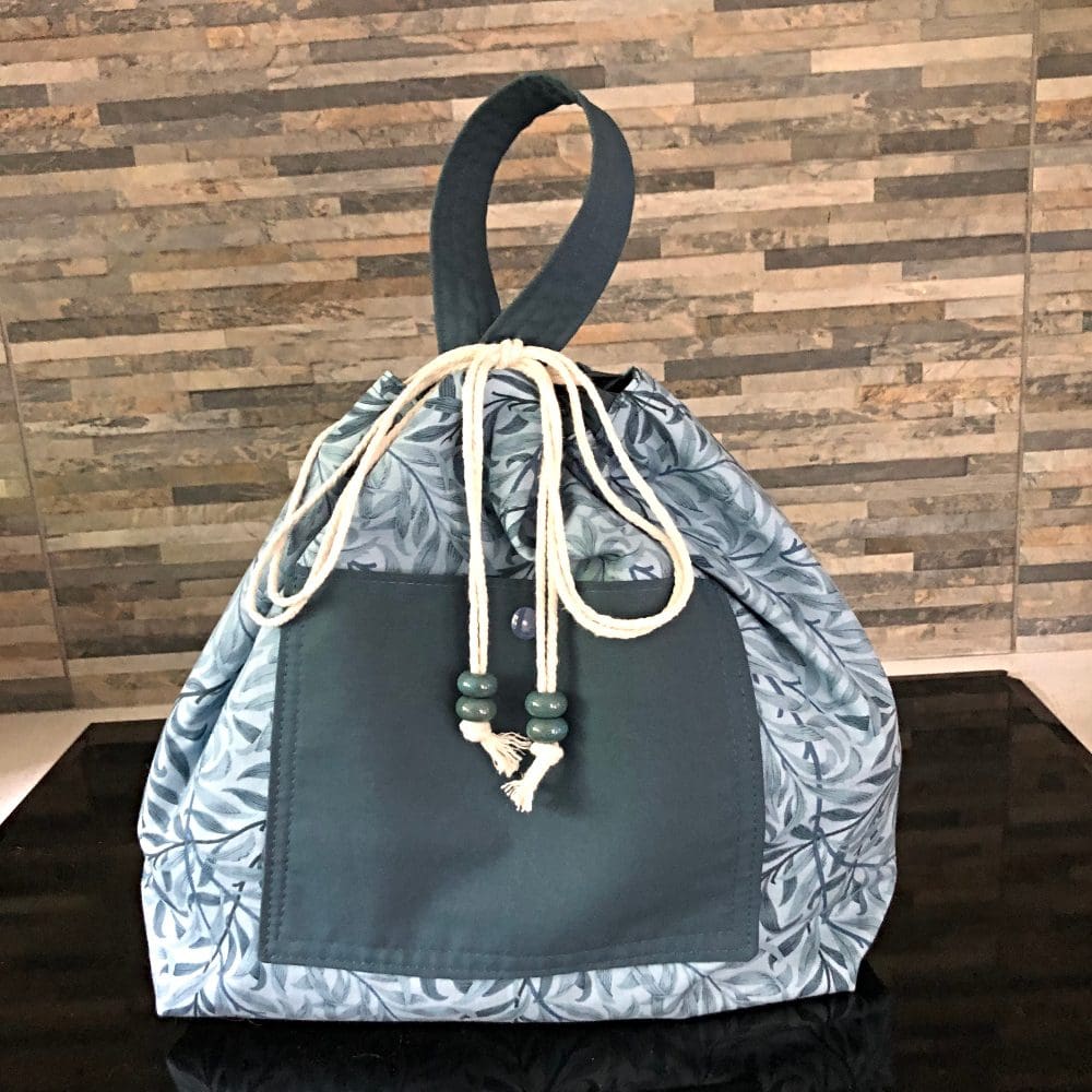 Closed and tied view of an extra large, drawstring bucket bag in William Morris Willows in shades of blue, with a contrasting snap pocket and handle. Closing with a chunky blue ceramic bead, this is standing on a black surface