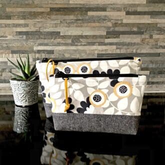 A pair of zipped bags using a cream, grey and mustard yellow fabric in an abstract floral design with a black zip and faux suede pull, standing on a shiny black surface with small pot plant