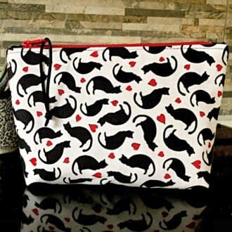 A medium sized zipped bag in white fabric printed with black cats and tiny red hearts standing on a shiny black surface. it has a red zip and lining.