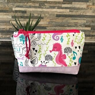 A small sized zipped bag in a pink and lilac seahorse designed fabric. The bag has a lilac linen base and lining, closing with a bright pink zip and pull faux suede pull. The bag stands on a shiny black surface.