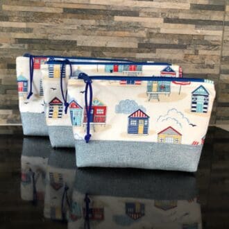 A set of three zipped pouches in white fabric with red, white and blue seaside hut design with royal blue zip and faux suede and dusky blue lining on shiny black surface.
