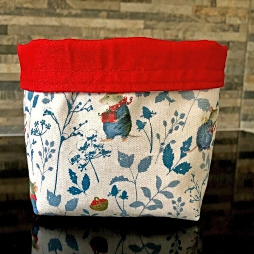 Reversible fabric pots in blue and red winter mice fabric and red lining with a flowering Christmas cactus inside on brick background