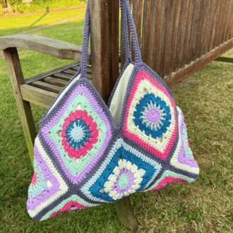 a beach bag made of granny squares in pink, teal, lilac and mint. It's hanging on a garden bench.