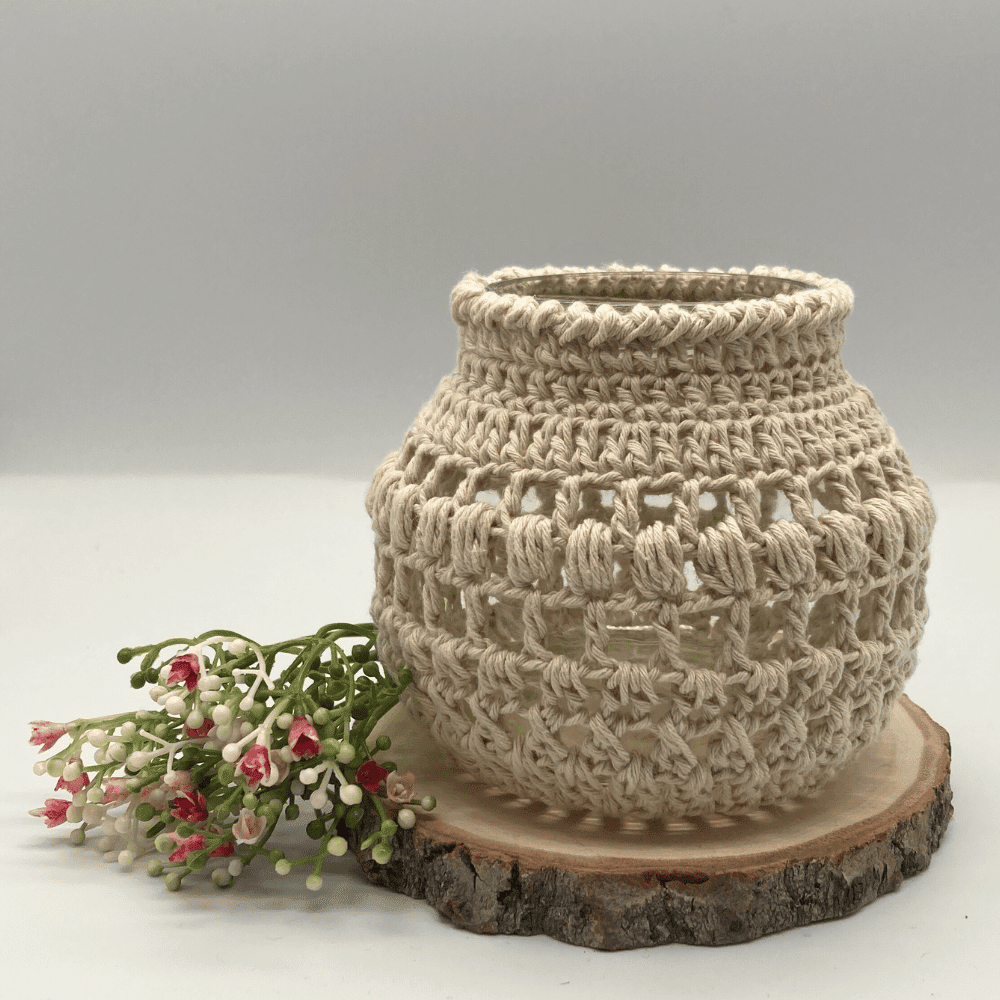 a round globe shaped jar with a cream crochet cover. It's sitting on a slice of wood and on the side are some small flowers