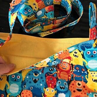 Adjustable cross body style mastectomy drain bag in Makowers colourful Aliens and bright yellow lining, on a reflective black surface