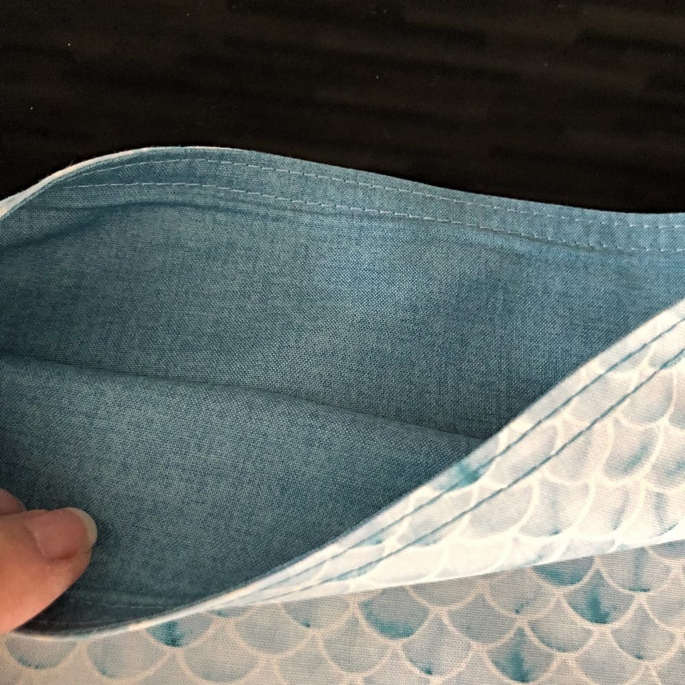 Adjustable cross body style mastectomy drain bag in Sanibel's Fish Scales fabric in shades of teal and cream with a pale blue linen-look cotton lining, on a reflective black surface
