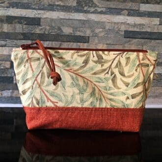 A small sized zipped bag in William Morris Willow fabric in green with a rust brown cotton base, lining, zip and faux suede pull. Standing on a reflective black surface.