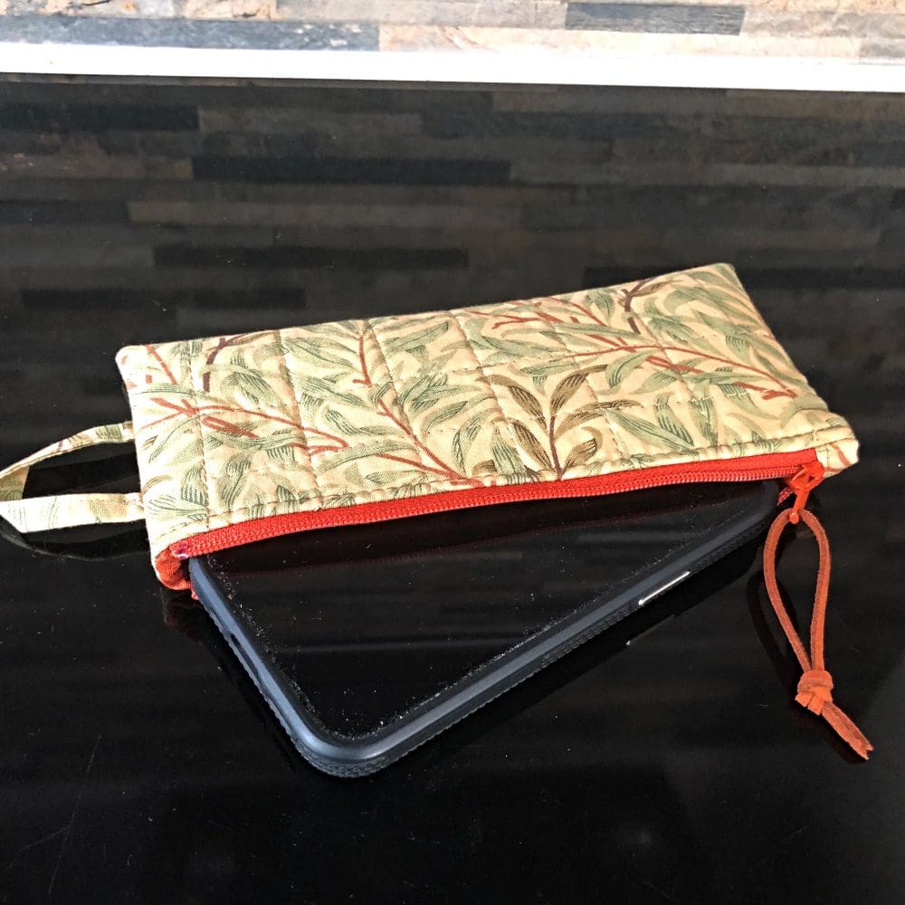 A small quilted phone wallet, shown open and holding an iphone12 in William Morris' Willow design in green on a black reflective black surface. Contrasting rust brown zip, faux suede pull and lining.