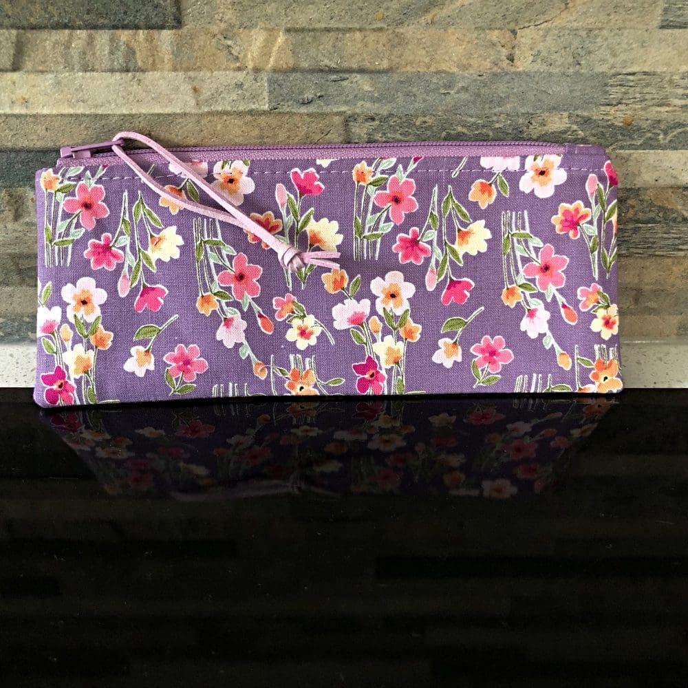 A zipped pencil case in lilac floral design with faux suede pull, choice of pale lilac zip and pink lining on shiny black surface.