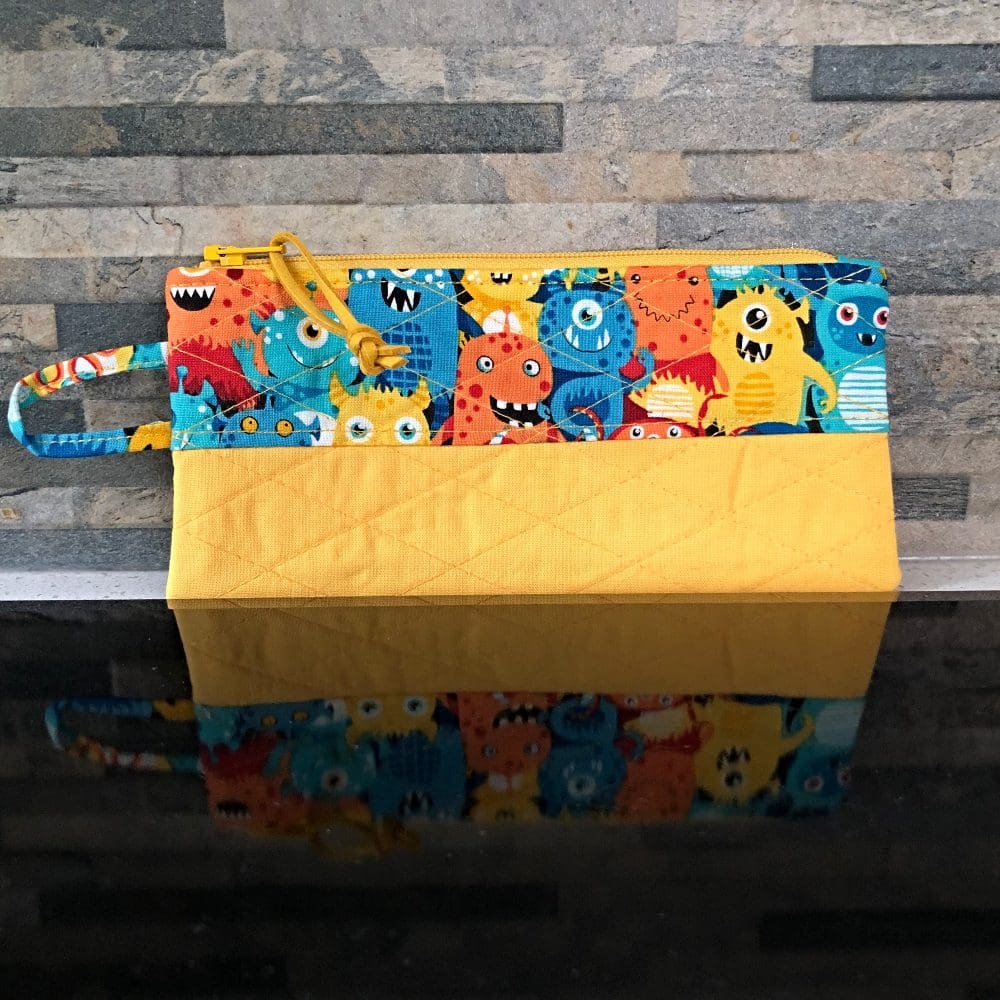 A small quilted phone wallet, in cute and colourful Aliens fabric. A bright yellow base, lining, zip and faux suede pull on reflective black surface