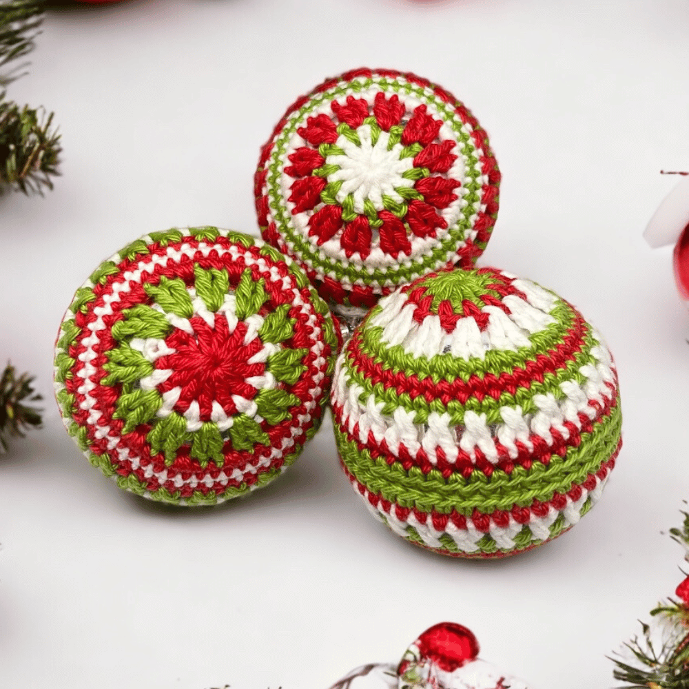 3 baubles enclosed in red white and green crochet. In the back ground there are red baubles and pine branches