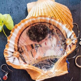 Gilded decoupage scallop shell trinket dish featuring Klimt's The Kiss, resting on a dark table surrounded by jewellery