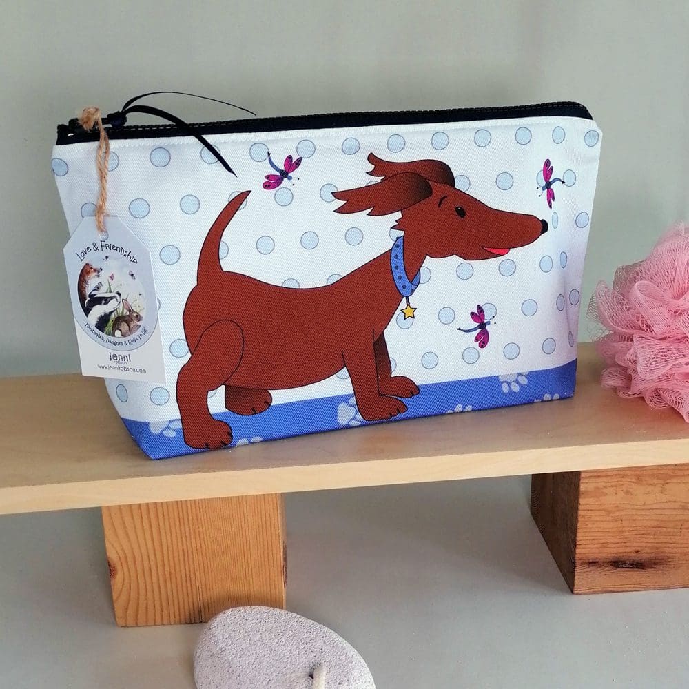 large cotton toiletries bag featuring a happy little brown dog in a blue collar standing against a blue spotty wall, surrounded by pink and blue dragonflies. The reverse of the washbag is a bright blue and there are paw prints on the base