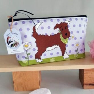 A large cotton handmade washbag featuring a background of pale purple spots, paw prints on the base and a cheeky brown dog with white paws, tummy and tail end, caught in the act of taking a white daisy from the garden. The illustration is on the front of the bag with a green base covered in paw prints.
