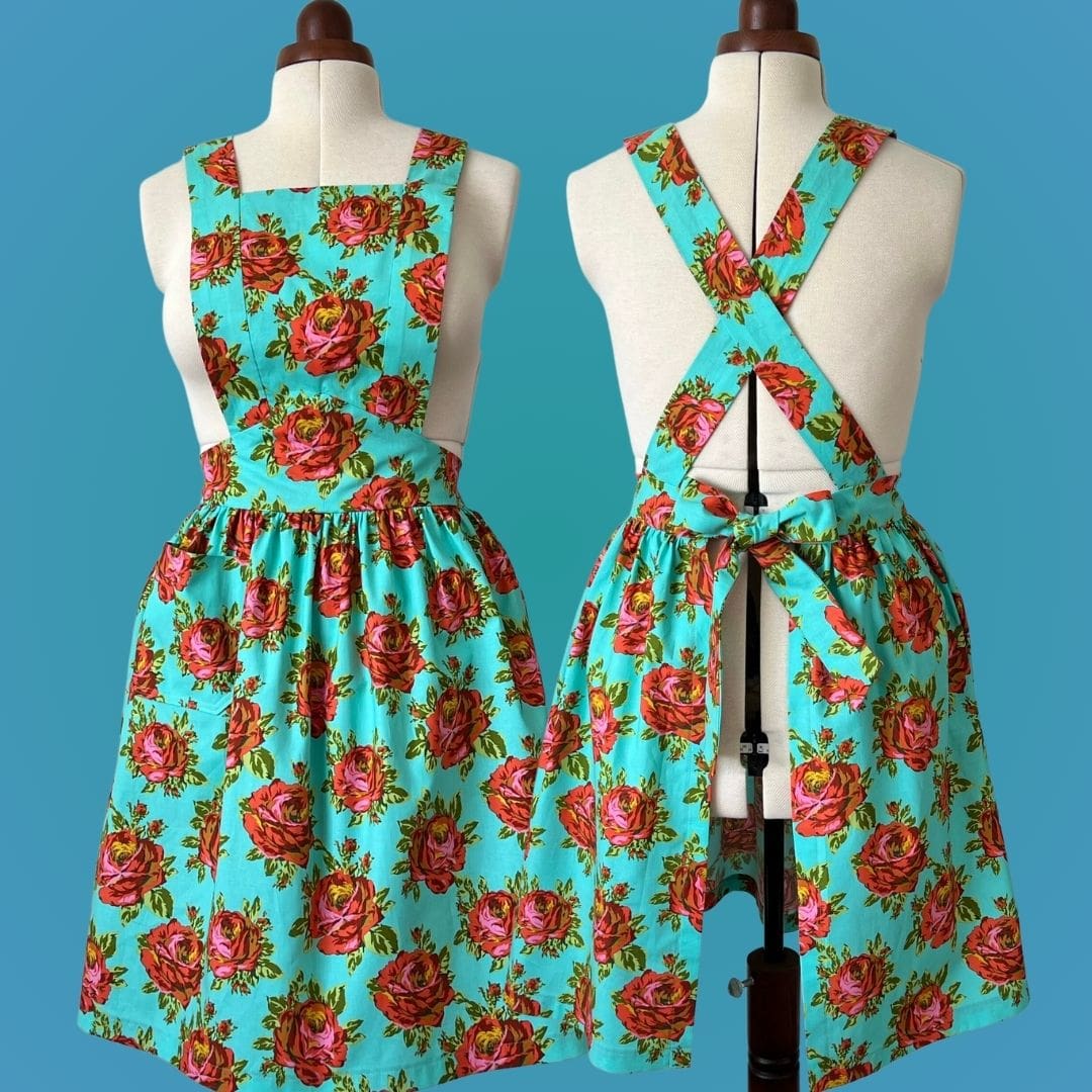 Two 1940s style aprons displayed side by side on vintage style mannequins, showing the front and back view pod the apron. Apron is made of a blue cotton fabric with a large red floral pattern.