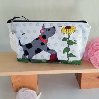 lA large cotton handmade toiletries bag featuring a background of pale blue spots, paw prints on the green base and a happy grey dog with large black spots balancing on a flower pot, watching the bees around a sunflower. The illustration is on the front of the bag with a pale blue cotton back.
