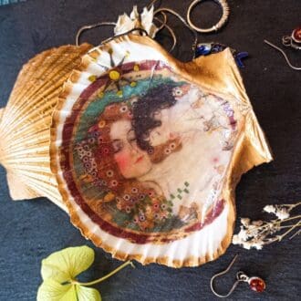 Gilded decoupaged scallop shell trinket dish with Klimt's Mother and Child design on a dark table surrounded by small flowers and jewellery