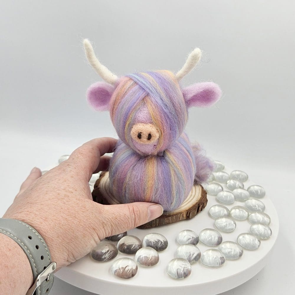 hand holding a Pastel coloured needle felted highland cow on wood slice.