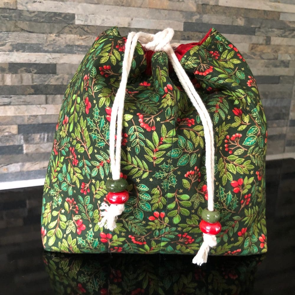 Medium sized quilted drawstring bag in a Christmas holly & berry fabric on a dark green background with red lining. Closes using a natural cord secured by chunky green and red ceramic beads.