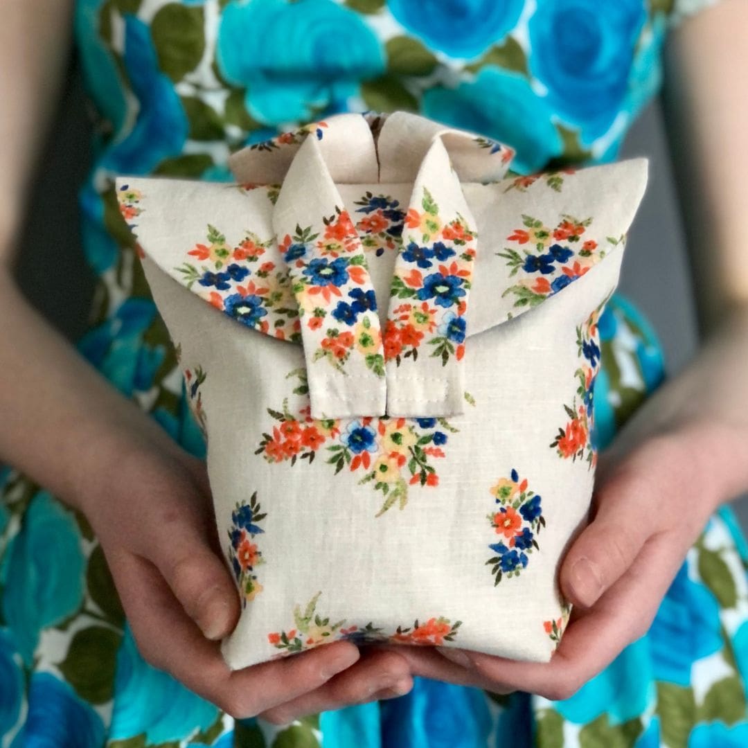 cream floral linen bag with two short straps held in the hands in front of a lady wearing a blue floral dress