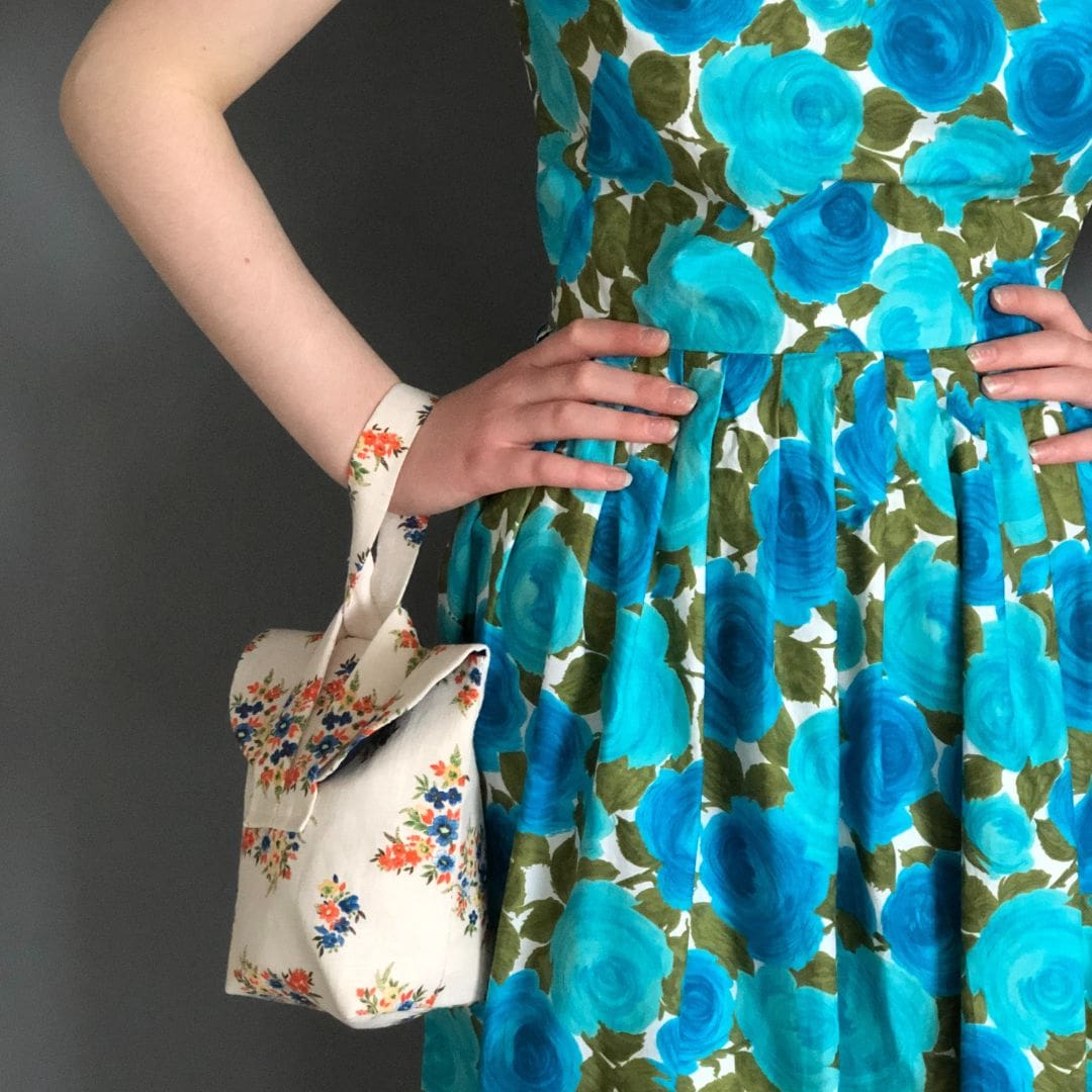 floral cream linen short strap bag hangs from the wrist of a lady wearing a blue floral dress