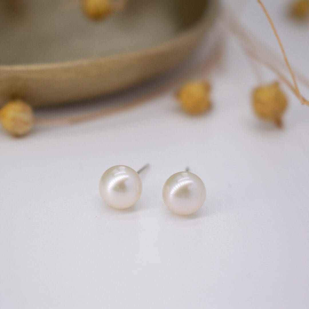 ivory pearl stud earrings on a white background with small brass dish and dried flowers