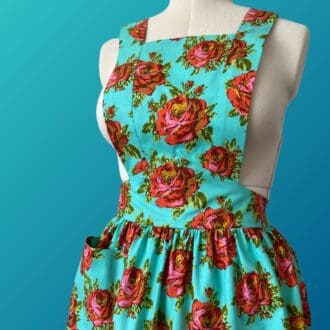 Front view of a 1940s style full bib apron, made from a vivid blue cotton fabric with a large red roses pattern. The apron is displayed on a vintage style mannequin.