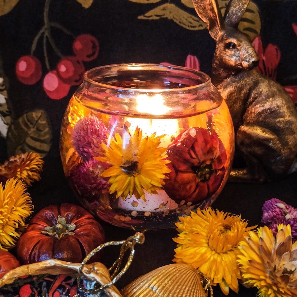 Autumn scented soy and gel wax candle decorated with brightly coloured dried flowers and pumpkins is lit and glows dramatically against a dark floral background next to a bronze hair