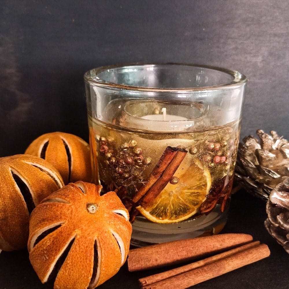 Orange spice soy and gel wax luxury tumbler with orange slices, cinnamon sticks, golden berries, star anise and crushed gold crystal rests on a dark background next to whole dried oranges and cinnamon sticks.