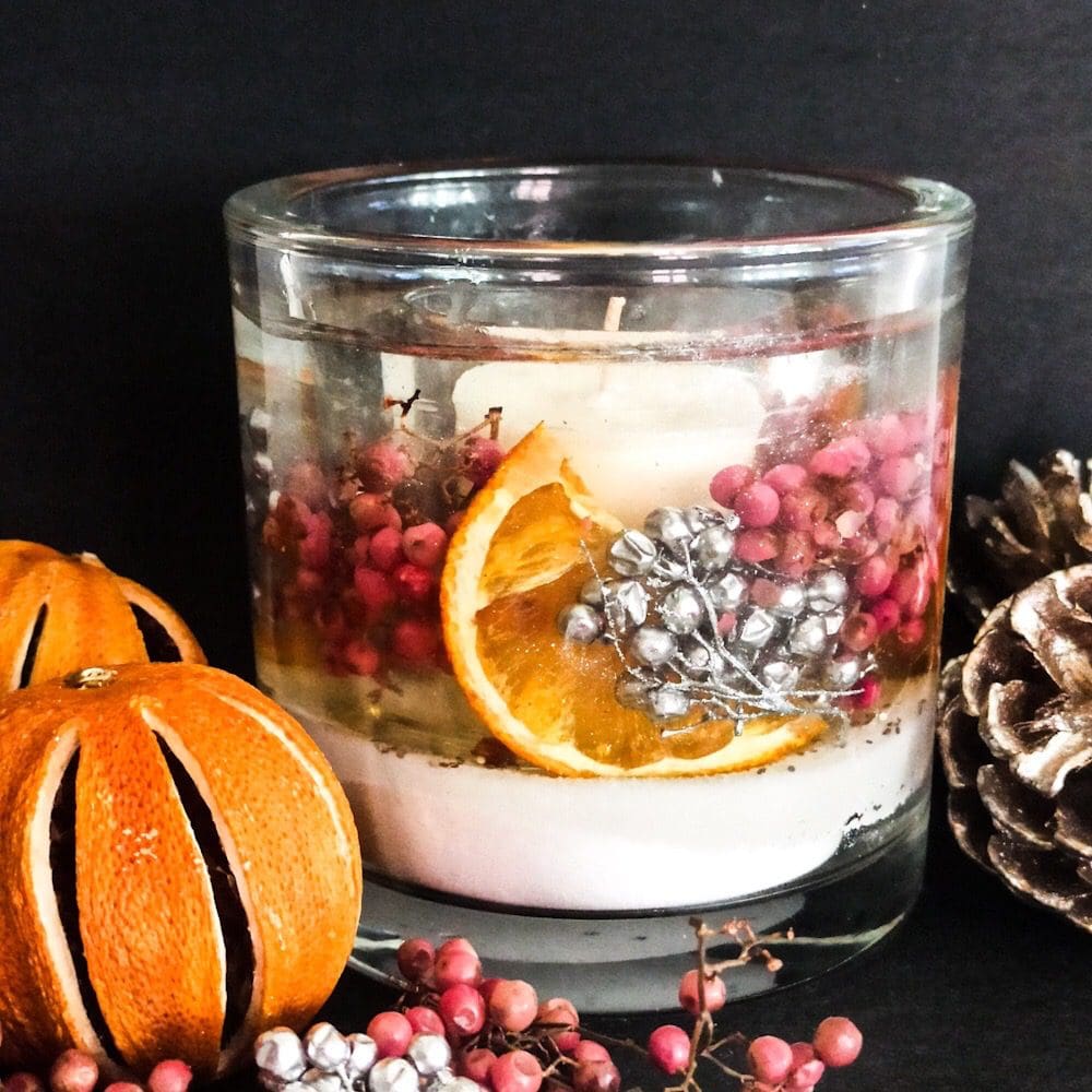 Sparkling cranberry soy and gel wax tumbler luxury scented candle containing dried orange slices and pink and silver berries is placed on a dark background surrounded by dried oranges and pink and silver berries