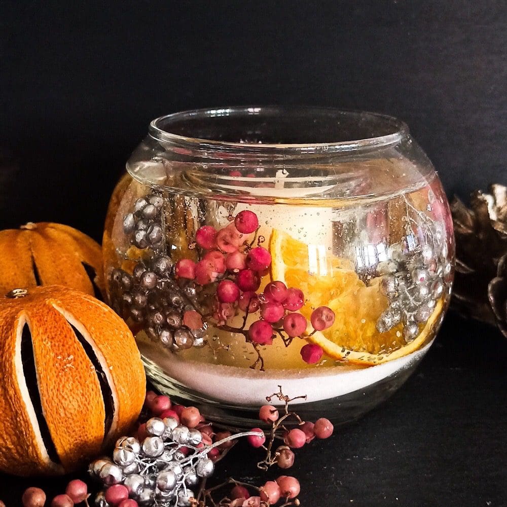 Sparkling cranberry soy and gel wax fishbowl scented candle with dried orange, pink and silver berries on a dark background, next to dried oranges and a scattering of pink and silver berries