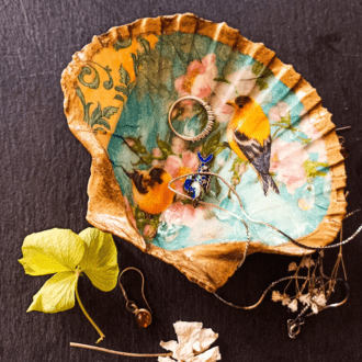 Gilded decoupage scallop shell trinket dish featuring two yellow birds against blue sky and cherry blossom rests on a dark surface with jewellery in and around the dish