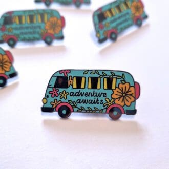 A close up of a turquoise, campervan shaped, enamel pin badge with orange and pink flowers and the words 'Adventure Awaits' in the centre of the motorhome design.