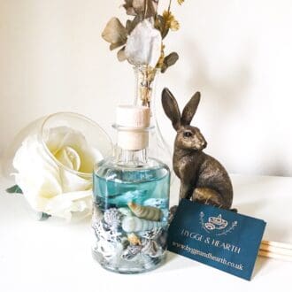 Blue sea luxury ocean reed diffuser with blue fragrance oil and shells is on a white background, next to a white rose, dried flowers, reeds, hygge and hearth business card and a bronze hare